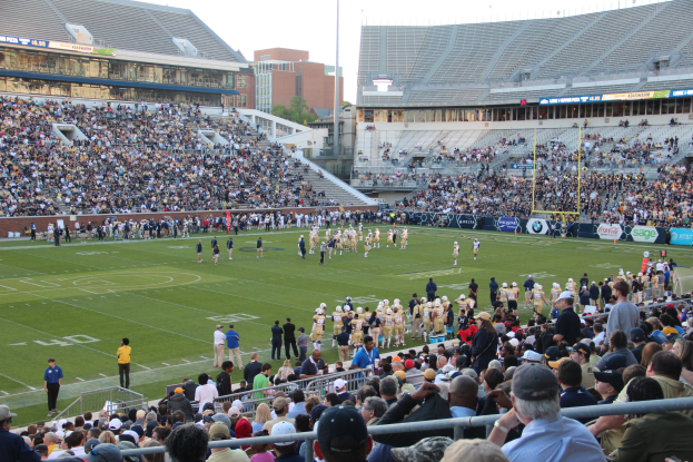 Ein Fußballspiel wird in einem Stadion mit Spielern auf dem Feld und Zuschauern in den Rängen gespielt, umgeben von Gittern, Texttafeln, Pfosten, Lampen, Bäumen und Gebäuden unter einem klaren blauen Himmel.