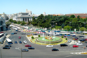 Eine belebte Stadtstraße mit verschiedenen Fahrzeugen, einem zentralen Brunnen, Laternen, Bäumen, Gebäuden und einem klaren blauen Himmel.