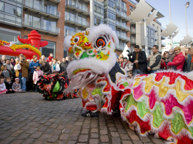 Ein lebhaftes chinesisches Neujahrsfest in Amsterdam mit einem Löwen tanzen im Vordergrund und einer Menge Menschen drumherum, Gebäuden und Laternenmasten im Hintergrund unter einem klaren blauen Himmel, während einige der Anwesenden den Moment mit Kameras festhalten.
