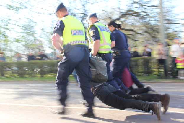 Eine Gruppe von Polizisten schiebt einen Mann die Straße entlang, wobei der Mann eine Tasche trägt und die Beamten Mützen aufhaben, vor einem unscharfen Hintergrund aus Bäumen, Pflanzen und einem Pfahl.