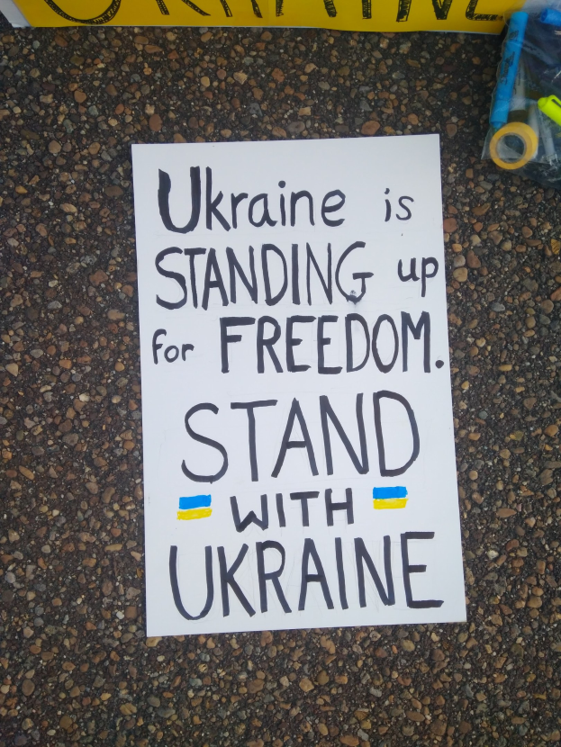 Ein weißer Zettel mit der Aufschrift "Ukraine steht für Freiheit, steht mit Ukraine" liegt vor einer gelben Fahne auf dem Boden, rechts daneben eine Plastikabdeckung mit verschiedenen Gegenständen.