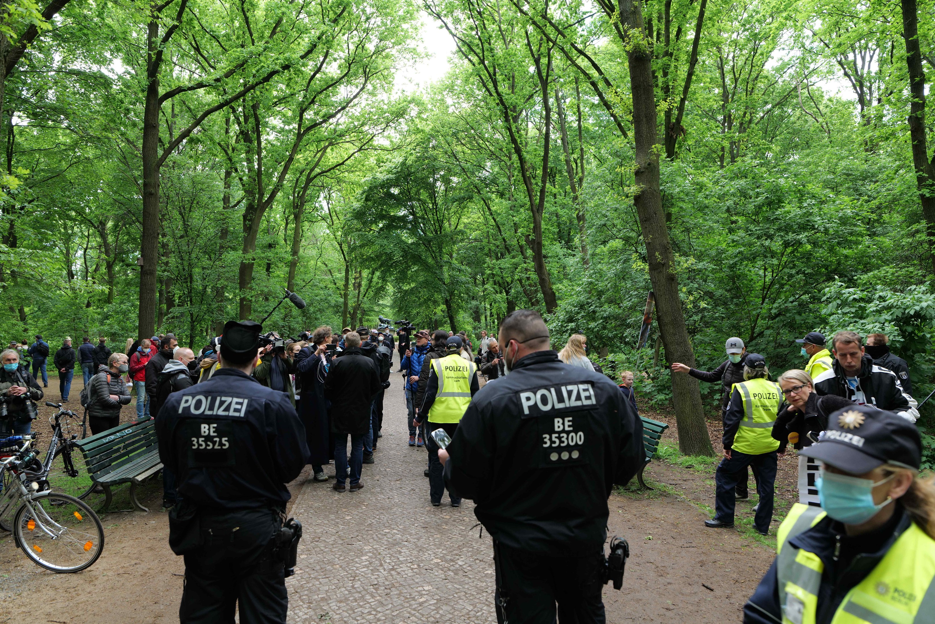 Polizisten vor einer Menge bei einer Anti-Terror-Demonstration in Berlin, mit Fahrrädern und einer Bank im Vordergrund und Bäumen im Hintergrund.