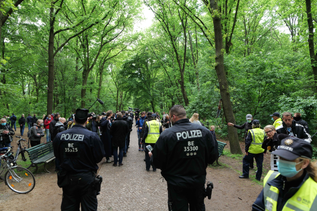 Polizisten vor einer Menge bei einer Anti-Terror-Demonstration in Berlin, mit Fahrrädern und einer Bank im Vordergrund und Bäumen im Hintergrund.