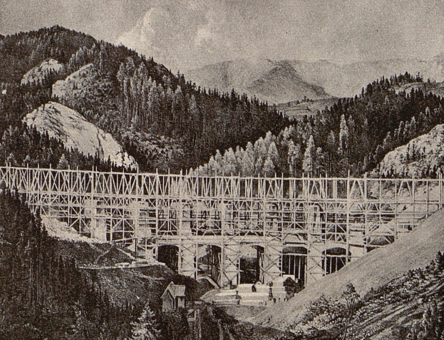 Schwarz-weiß-Foto einer Kabelbrücke im Bau, die ein bergiges Gelände mit Bäumen, Hügeln und einem bewölkten Himmel überspannt.