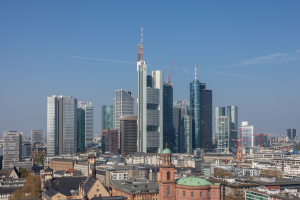 Die Frankfurter Skyline von einem Hügel aus betrachtet, mit einem prominenten Wolkenkratzer in der Mitte umgeben von anderen Gebäuden und Bäumen, mit dem Himmel im Hintergrund.