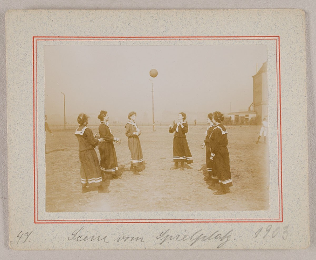 Eine Gruppe von Frauen, die Fußball auf einem Feld mit einem Ball in der Mitte, Pfosten und Gebäuden im Hintergrund und Text unten auf dem Bild spielen.