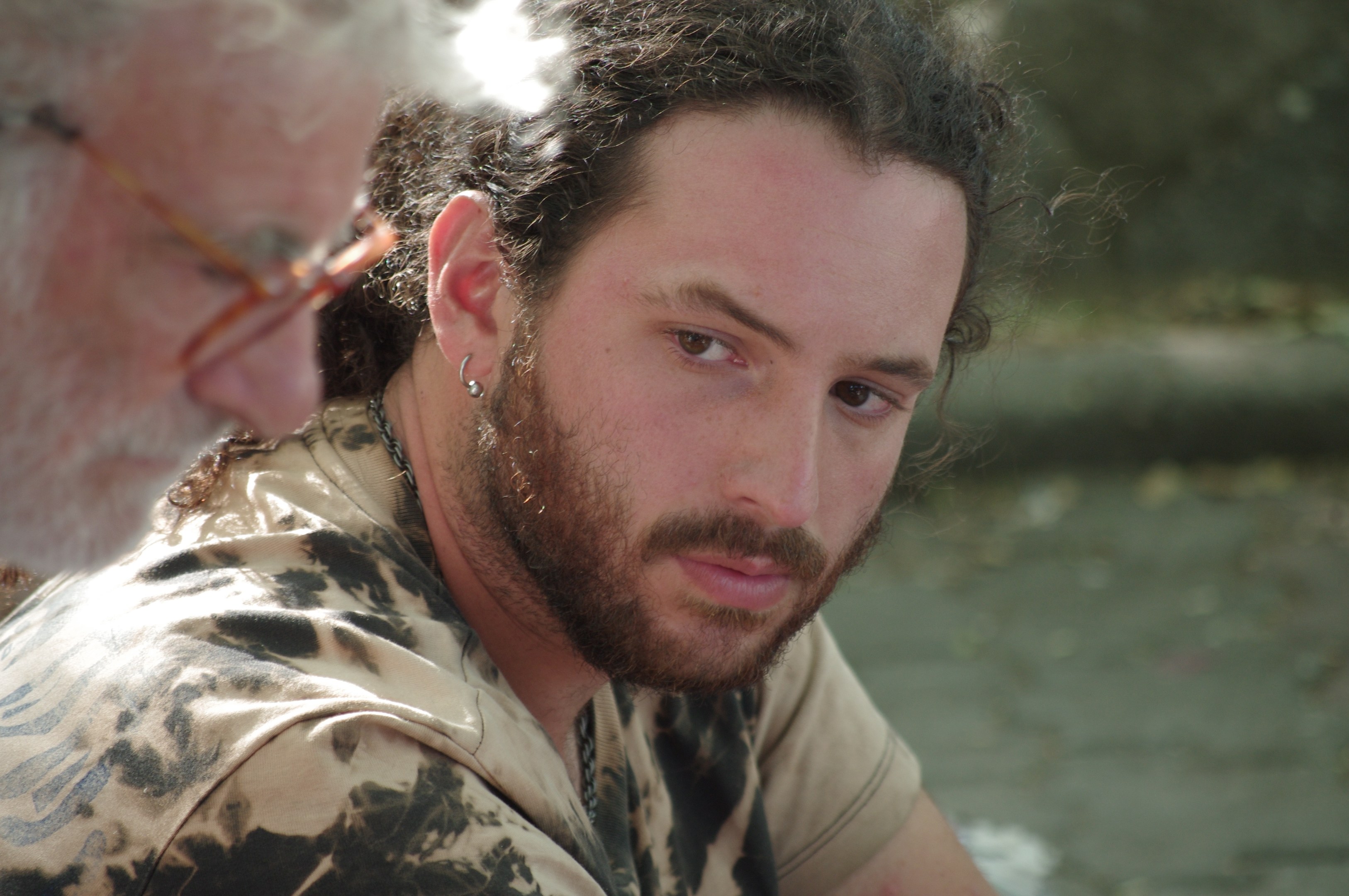 Zwei Männer sitzen nebeneinander am Wasser, einer trägt eine Brille, mit einem sanft unscharfen Hintergrund.
