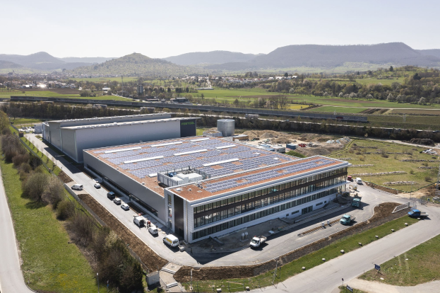 Luftaufnahme eines großen Industriegebäudes mit Solarpaneelen auf dem Dach, umgeben von Fahrzeugen, Bäumen, Gras, Strommasten und Bergen in der Ferne.