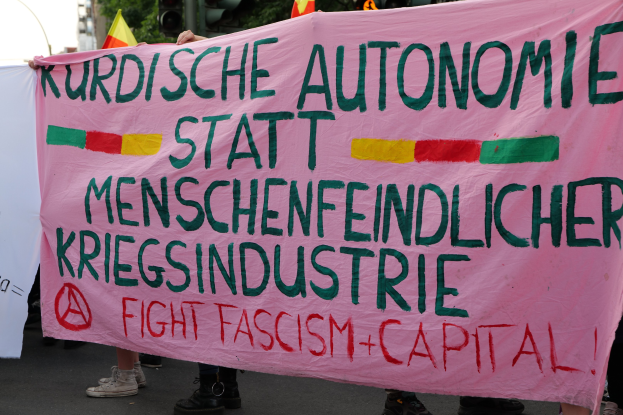 Eine Gruppe von Menschen steht auf einer Straße und hält ein pinkes Banner mit der Aufschrift "Kurdische Autonomie Statt Menschenfeindlicher Kriegsindustrie Fight Fascism Capital", mit Verkehrszeichen, Fahnen, Bäumen, Gebäuden und einem klaren blauen Himmel im Hintergrund.