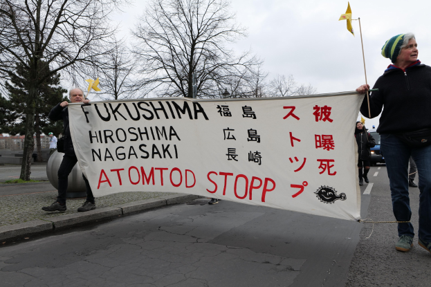 Ein Mann hält ein Banner mit der Aufschrift "Fukushima Hiroshima Nagasaki Atomod Stop" und geht durch eine Straße, umgeben von Menschen mit Fahnen, Bäumen, Gebäuden und einem klaren blauen Himmel im Hintergrund.