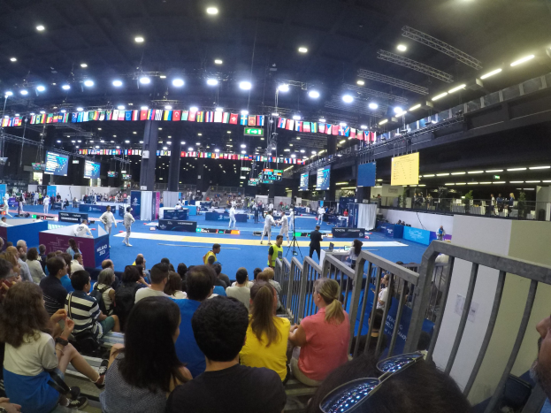 Eine große Menschenmenge sitzt und steht in einer Indoor-Sportarena mit Bildschirmen, Flaggen und Deckenleuchten im Hintergrund.