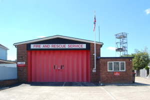 Feuerwehr- und Rettungsdienstgebäude mit roter Tür, Fenstern, einem Namensschild, einem Flaggenmast mit Flagge, einem Metallturm, einem Zaun, einer Baumgruppe und einem bewölkten Himmel.