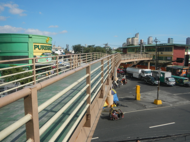 Eine Fußgängerbrücke mit Geländern auf beiden Seiten überspannt eine vielbefahrene Straße voller Autos und Lastwagen, mit Gebäuden, Bäumen, Pfählen, Drähten und einem klaren blauen Himmel im Hintergrund.