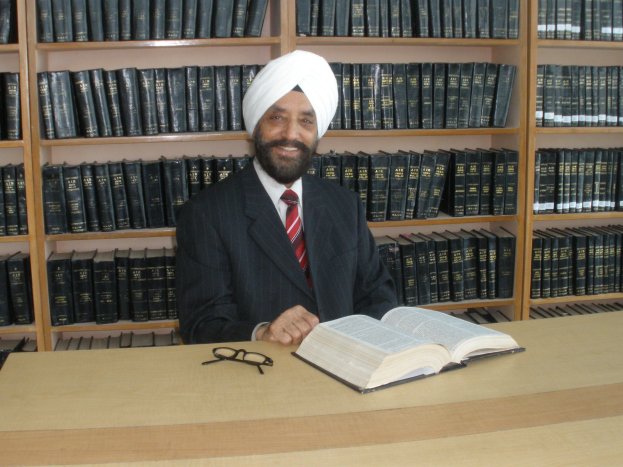 Ein Mann in Anzug und Krawatte sitzt an einem Schreibtisch mit einem Buch und einer Brille, umgeben von Bücherregalen voller Bücher.