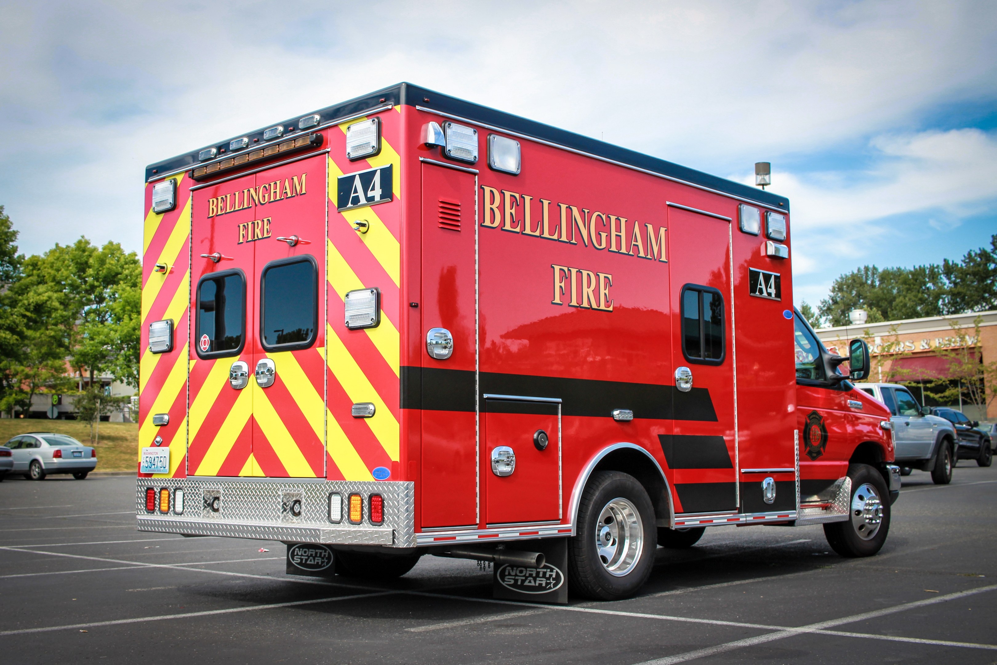 Ein Rettungswagen des Bellingham Fire Department auf einem Parkplatz mit Bäumen und Gebäuden geparkt, unter einem klaren blauen Himmel.
