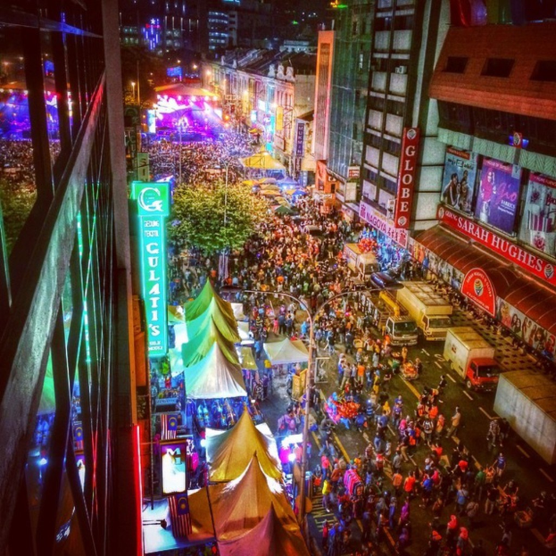 Ein volles Karnevalsszenario mit Zelten, Ständen, Gebäuden und Bannern, die die Straße säumen, mit beleuchteten Strukturen im Hintergrund.