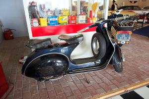 Ein schwarzer und weißer Roller steht vor einer Tankstelle, mit einem roten Objekt und einem Feuerlöscher auf der linken Seite und mehreren Fahrzeugen, einer Person und Brettern an der Wand im Hintergrund.