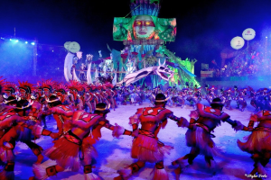Eine Gruppe von Menschen in farbenfrohen Kostümen tanzt auf einer Bühne vor einem sitzenden Publikum, mit Lichtern, Stangen und Ballons, und einem Bildschirm mit einer Person im Hintergrund, während des Karnevals in Rio de Janeiro 2014.