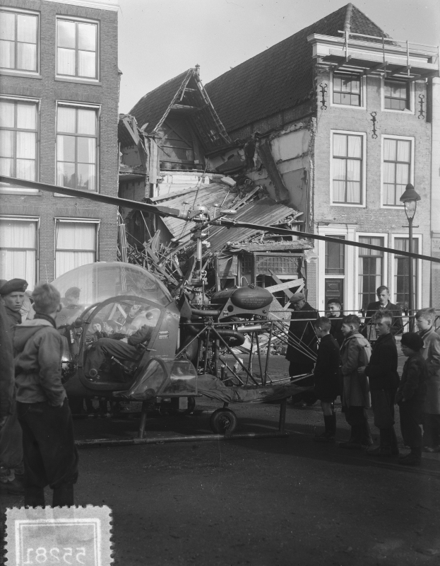 Schwarzes und weißes Bild eines kleinen Flugzeugs, das in ein Gebäude gekracht ist, mit Menschen drumherum, einer Person sichtbar im Fahrzeug und einer Straßenlaterne in der Nähe.