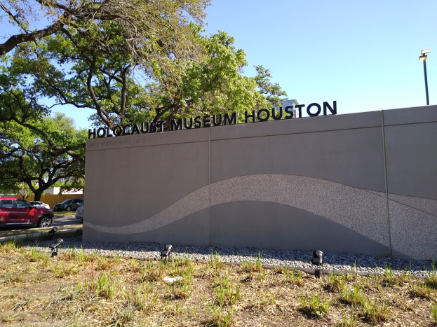 Außenansicht des Holocaust-Museums Houston mit eingravierter Textwand, Gedenksteine, Landschaftsgestaltung, Straße mit Fahrzeugen, Straßenmast und bewölktem Himmel.