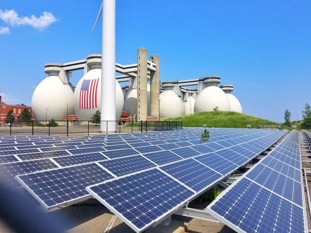 Solarpanels auf dem Dach eines Gebäudes mit einer Flagge, umgeben von einem Zaun, Grünfläche, Bäumen und anderen Strukturen, vor einem bewölkten Himmel.