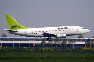Ein AirBaltic Airbus A320-200-Flugzeug startet am Frankfurter Airport, umgeben von grünem Gras, bunten Blumen und verschiedenen Hintergrundelementen wie einem Gebäude, Bäumen, Pfählen und Fahrzeugen.