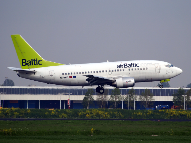 Ein AirBaltic Airbus A320-200-Flugzeug startet am Frankfurter Airport, umgeben von grünem Gras, bunten Blumen und verschiedenen Hintergrundelementen wie einem Gebäude, Bäumen, Pfählen und Fahrzeugen.