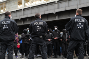 Eine Gruppe von Polizisten in Uniform steht vor einer Menge von Menschen in schwarzen Uniformen und Masken, mit einer Brücke und einem Gebäude im Hintergrund, was auf eine städtische Umgebung während einer Demonstration hindeutet.