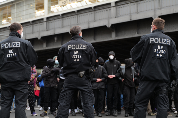 Eine Gruppe von Polizisten in Uniform steht vor einer Menge von Menschen in schwarzen Uniformen und Masken, mit einer Brücke und einem Gebäude im Hintergrund, was auf eine städtische Umgebung während einer Demonstration hindeutet.