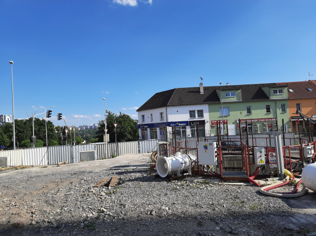 Baustelle mit freiliegenden Rohren und Geräten auf dem Boden, umgeben von Gebäuden, Laternen, Verkehrssignalen, einem Zaun, Bäumen und einem bewölkten Himmel.