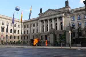 Das Reichstagsgebäude in Berlin, Deutschland, ein großes Gebäude mit zahlreichen Fenstern, umgeben von Straßen-Elementen, Fahrzeugen, Menschen und Flaggen, unter einem bewölkten Himmel.