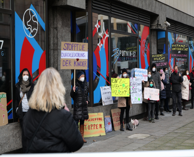 Menschen mit Masken und Schildern vor einem gläsernen Gebäude, mit Kameras und einem Hund, protestieren gegen eine deutsche Regierungspolitik.