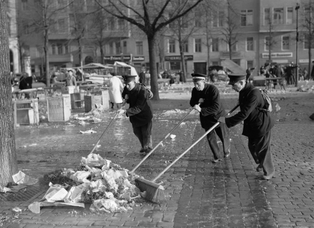 Drei Männer mit Mützen, die Müll auf einer Straße mit Bäumen, Gebäuden und Laternen in Schwarz-Weiß kehren.