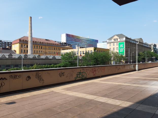 Stadtstraße mit Graffiti-beschmierten Wand, Bäumen, Straßenlaternen, Hinweisschild, Fahrzeugen, Gebäuden mit Fenstern, einem Schornstein und einem bewölkten Himmel.