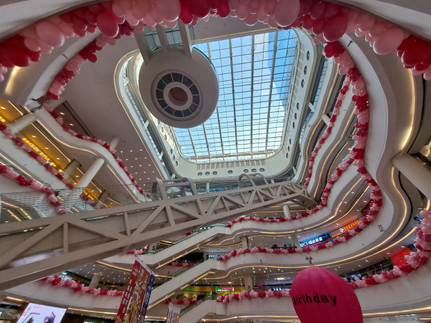 Ein Einkaufszentrum-Innenraum dekoriert für den Valentinstag mit pinken und weißen Ballons, Rolltreppen, Geländern, Texttafeln, Lichtern und einer Glasdecke.