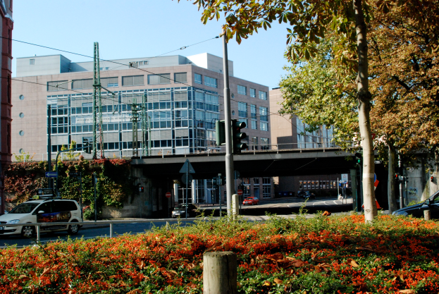 Eine Stadtstraße mit Autos, umgeben von blühenden Pflanzen, Bäumen, Polen, Verkehrsampeln, Schildern, einer Brücke, Gebäuden mit Fenstern und einem Himmel im Hintergrund.