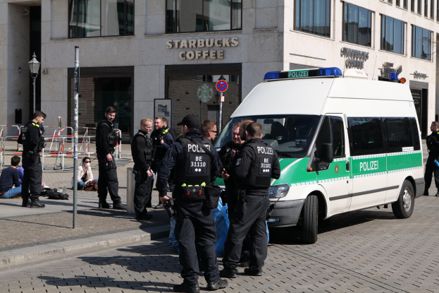 Eine Gruppe von Polizeibeamten steht vor einem Starbucks-Café, mit einem Van auf der rechten Seite und einigen Menschen auf der linken Seite und einem Gebäude mit Fenstern, einem Schild, einem Laternenpfahl und einem Zaun im Hintergrund.