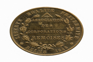 Goldmedaille mit der Aufschrift "Association des Corporations Remoises" auf weißem Hintergrund.