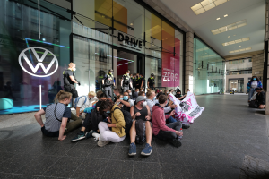 Menschen sitzen vor einem Volkswagen-Hndler auf dem Boden und halten ein Plakat waehrend einer Demonstration.