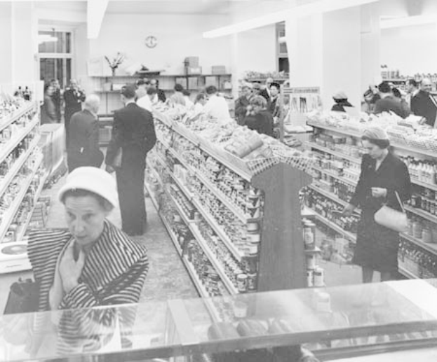 Schwarzes und weißes Foto von Menschen beim Einkaufen in einem Supermarkt mit Lebensmittelregalen, einige halten Taschen, eine Uhr an der Wand und Deckenleuchten.