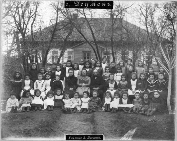 Schwarzes und weißes Foto einer Gruppe von Kindern, die vor einem Fensterbau posieren, umgeben von Bäumen und einem klaren Himmel, mit Text unten: "Jugoslawien, die erste Schule der Welt".