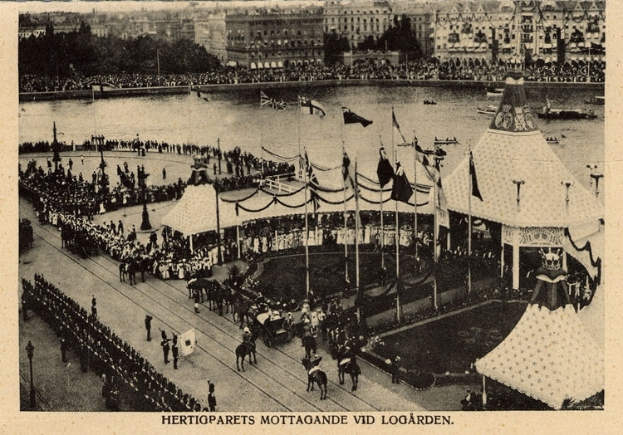 Ein altes Schwarz-Weiß-Foto einer Menge, die sich um einen Karneval mit Zelten, Fahnen, Stangen und Pferden im Vordergrund versammelt hat, und Gebäuden, Bäumen und Wasser im Hintergrund, mit Text am unteren Rand des Bildes.