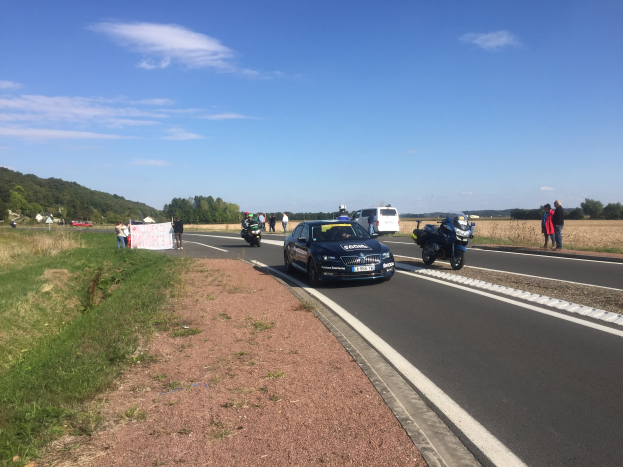 Eine Gruppe von Menschen steht neben einem Polizeiauto und einem Motorrad am Straßenrand, mit einem Banner und einem bewölkten Himmel im Hintergrund.