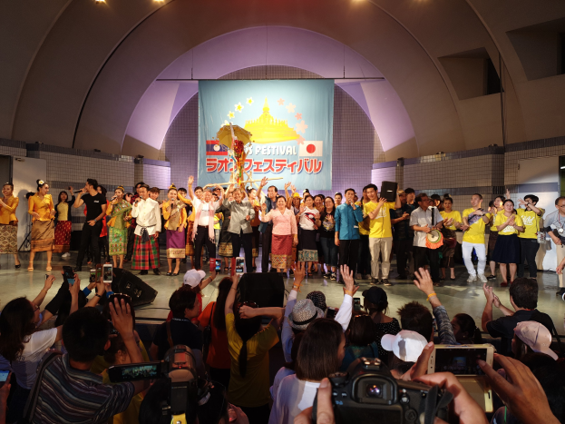 Eine Gruppe von Menschen auf einer Bühne mit Kameras, Handys und Musikinstrumenten während einer Festivaleröffnung mit Bannern, Text und Deckenlampen im Hintergrund.