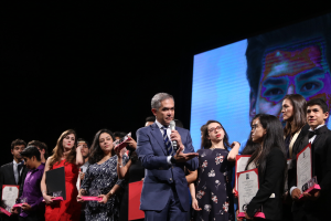 Ein Mann in Anzug und Krawatte hält ein Mikrofon auf der Bühne, umgeben von Menschen mit Auszeichnungen, mit einem Bildschirm im Hintergrund.