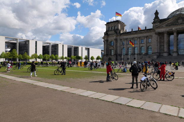 Eine Gruppe von Menschen, die Fahrräder vor dem Reichstaggebäude in Berlin, Deutschland fahren, mit dem Gebäude geschmückt mit Flaggen und Säulen, grasbewachsenem Boden, Bäumen im Hintergrund und einem bewölkten Himmel.