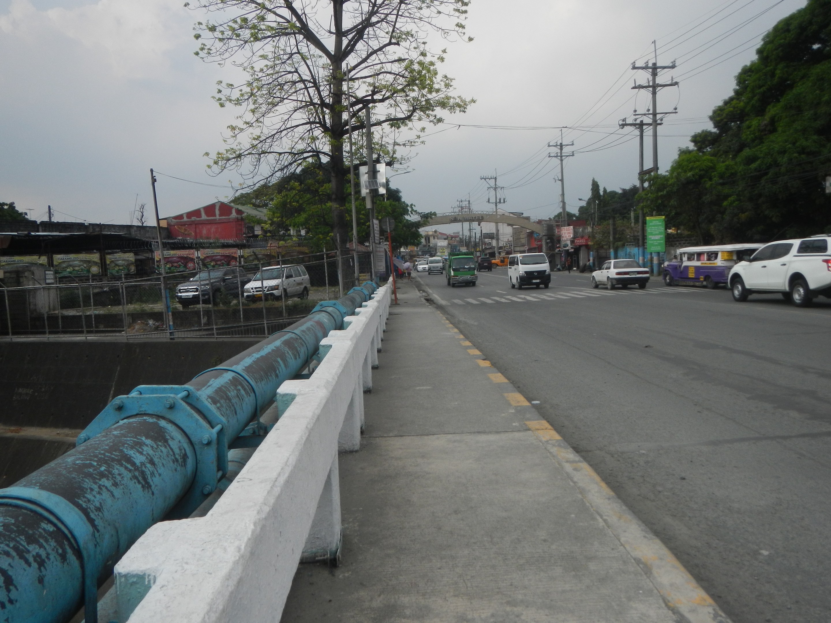 Stadtstraße mit fahrenden Autos, eine entfernte Brücke, Strommasten mit Drähten, Bäume, Gebäude und ein bewölkter Himmel, mit einem blauen Rohr an der Straßenseite.
