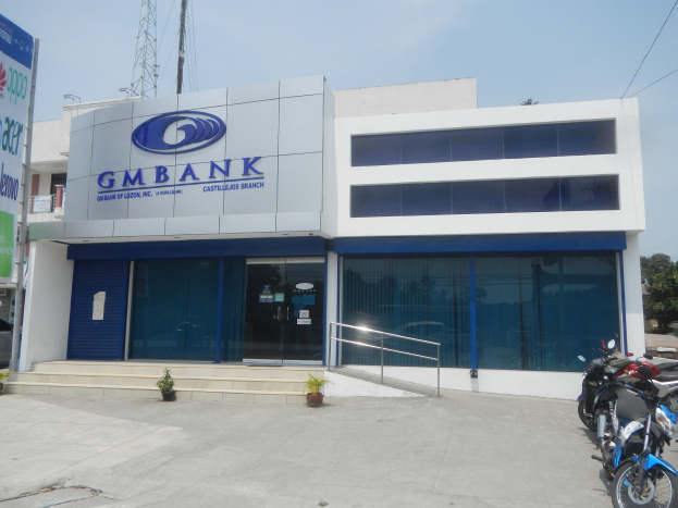 Außenansicht der Gmbank Bank in Hyderabad mit Glaswänden, Treppen mit Geländern, Topfpflanzen, Straßenfahrzeugen, einem Banner, Versorgungsleitungen, Bäumen und einem bewölkten Himmel.
