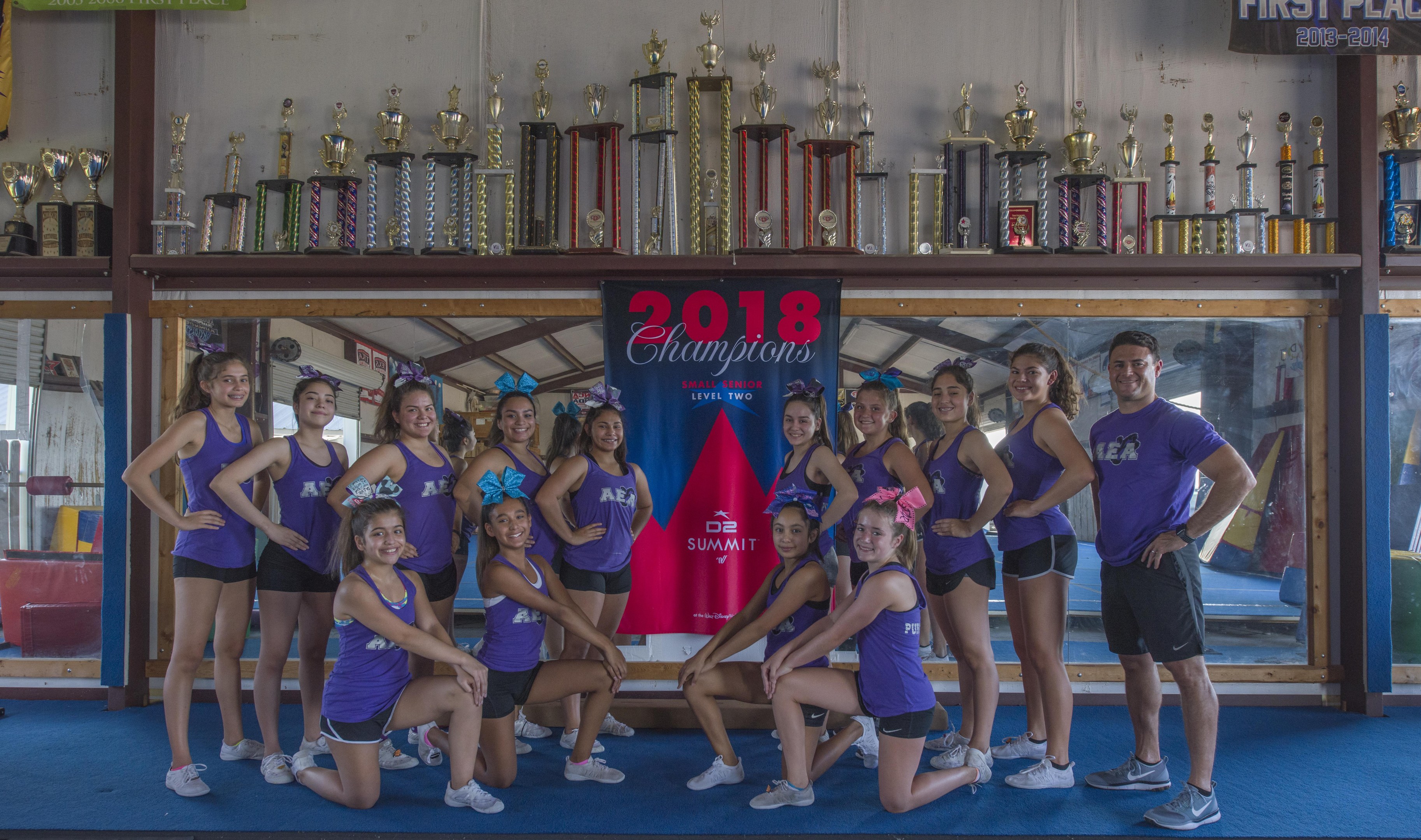 Eine Gruppe von Cheerleadern in lila T-Shirts und schwarzen Shorts posiert vor einem Trophy-Vitrinenregal mit einem Banner "2018 Summit Cheerleading Champions" im Hintergrund.