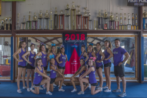 Eine Gruppe von Cheerleadern in lila T-Shirts und schwarzen Shorts posiert vor einem Trophy-Vitrinenregal mit einem Banner "2018 Summit Cheerleading Champions" im Hintergrund.